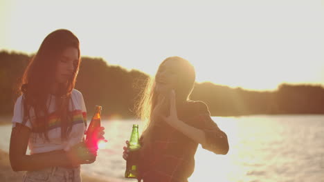 Two-female-students-with-perfect-young-bodies-are-dancing-in-short-t-shirts-with-beer-on-the-beach-party-at-sunset.-Their-long-hair-is-flying-on-the-wind.
